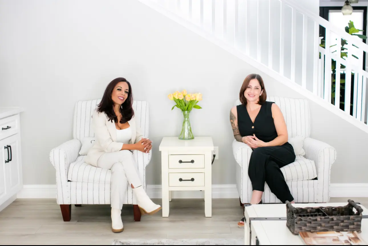 Two women sitting in chairs next to a table.